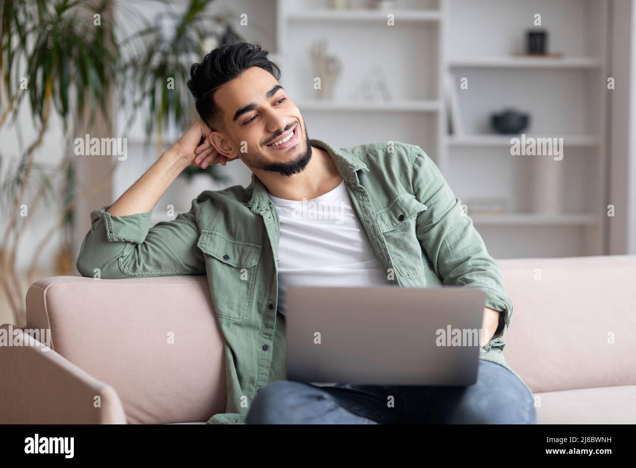 Jeune musulman souriant avec montre de barbe dans l'espace libre, assis sur un canapé avec ordinateur portable dans le salon intérieur Banque D'Images