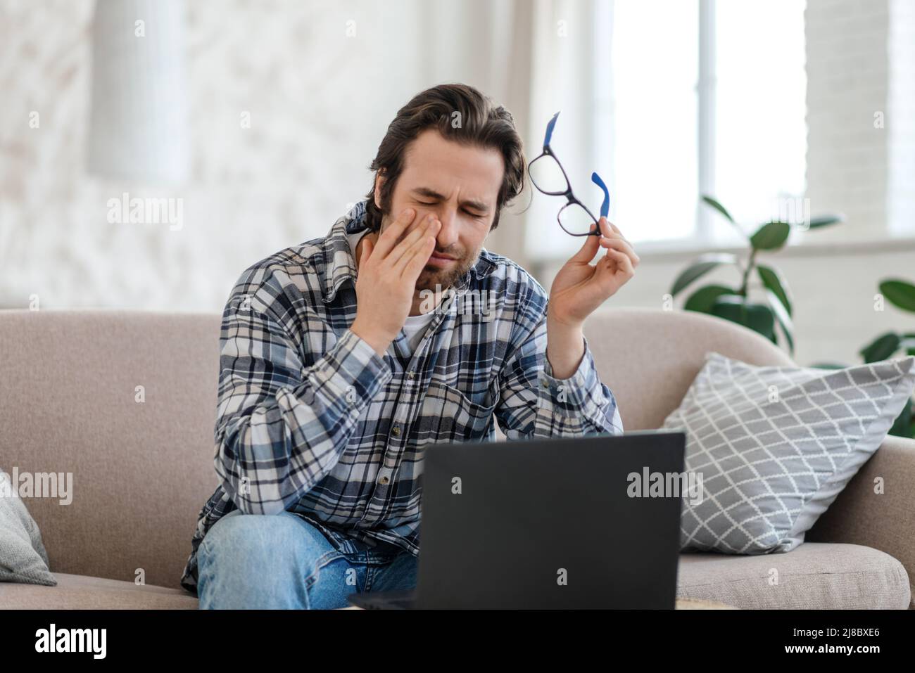 Triste millennium caucasien gars avec chaume s'assoit sur le canapé avec ordinateur portable prend des lunettes, souffre de maux de tête Banque D'Images