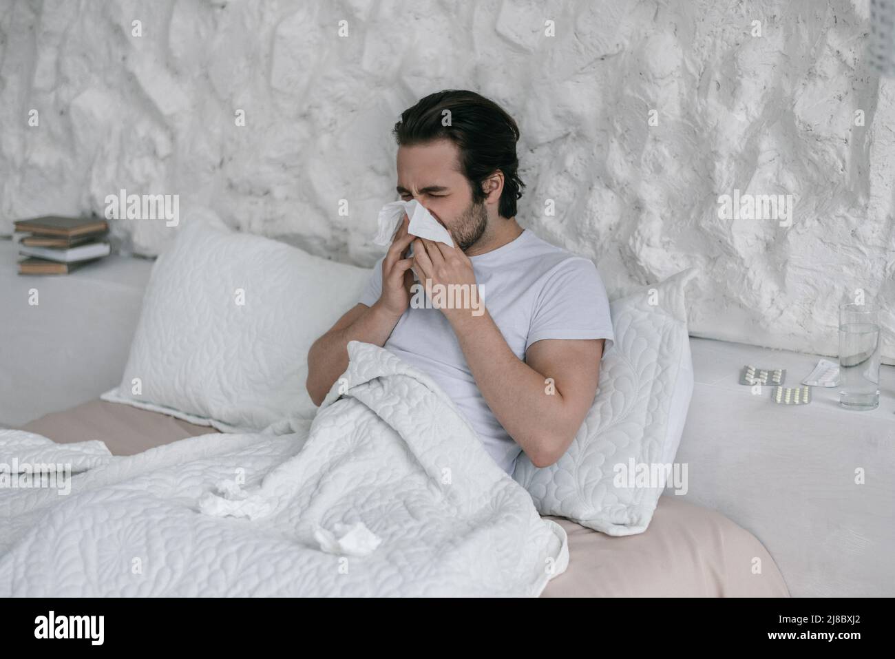 Un homme européen millénaire malheureux se trouve sur le lit, souffle le nez à la serviette dans l'intérieur blanc de la chambre Banque D'Images