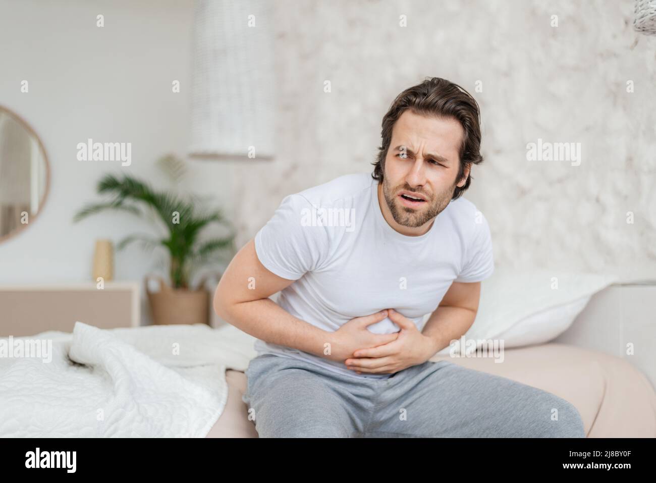 Mécontent millennal caucasien gars avec le repos de chaume, assis sur le lit blanc appuie ses mains à l'estomac Banque D'Images