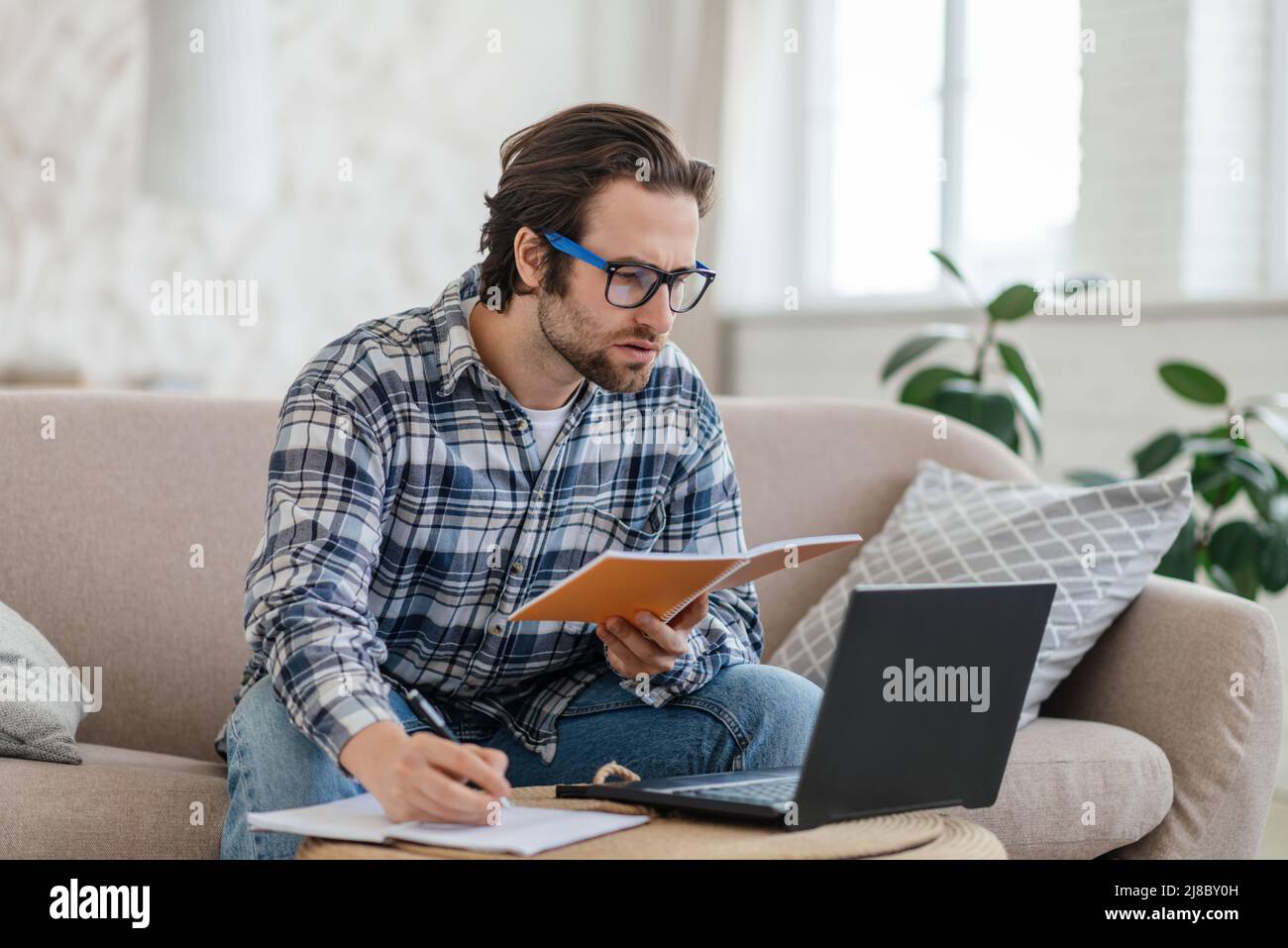 Homme caucasien millénaire occupé avec un chaume dans des lunettes est assis sur un canapé avec ordinateur portable et livre, recherche, prend des notes Banque D'Images