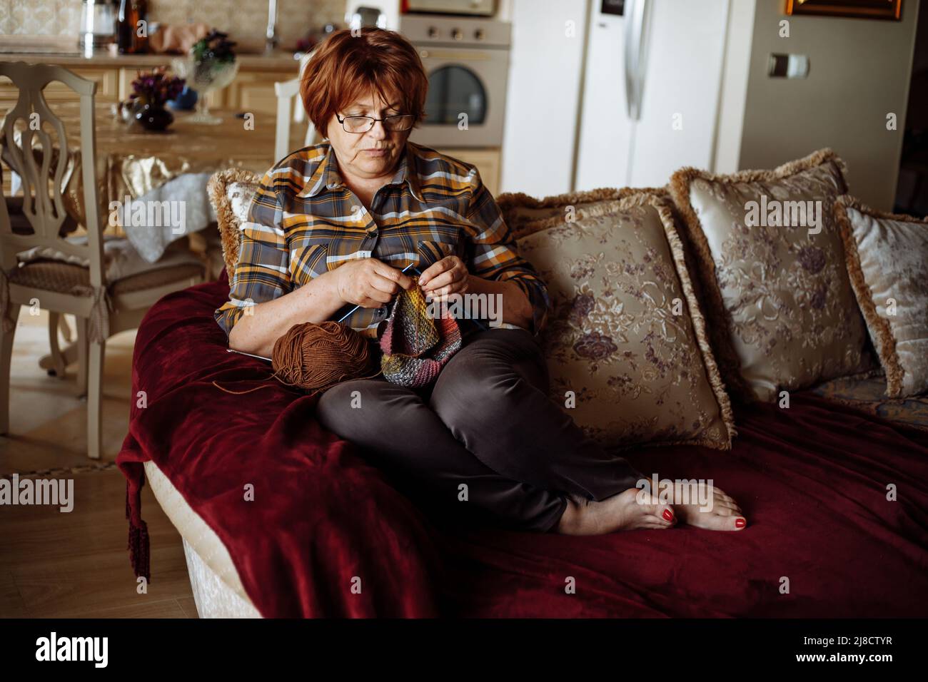 Femme mûre rouge cheveux dans des verres assis sur le canapé dans le salon et tricotage à l'aide d'aiguilles et de boule de fil. Maison de retraite de loisirs et fait à la main Banque D'Images