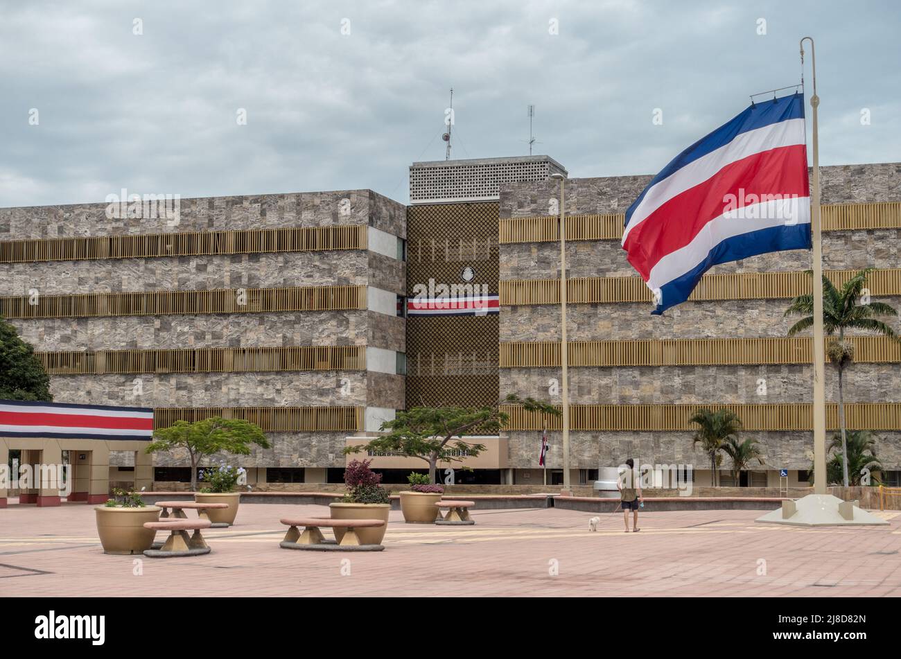 Homme marchant son chien sous un grand drapeau costaricain au système judiciaire de San José. Banque D'Images