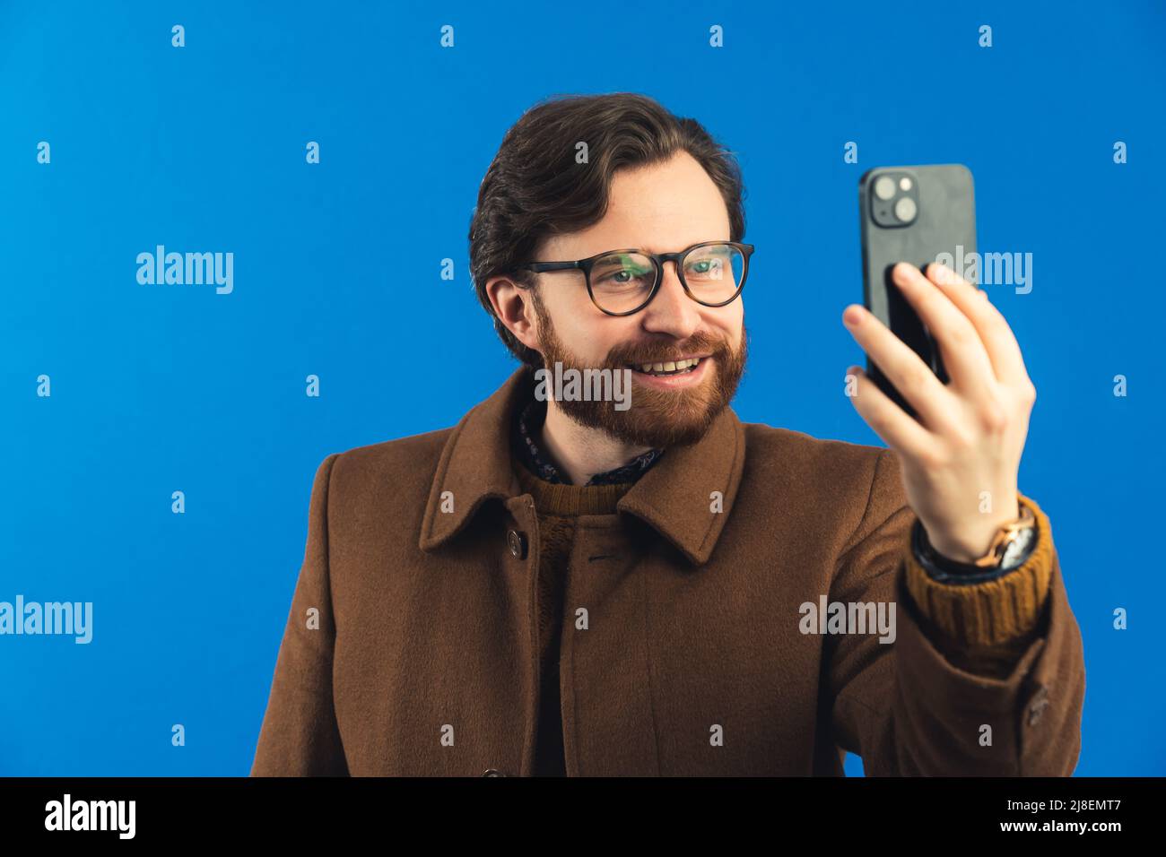 Studio de gros plan moyen tourné sur fond bleu. Homme heureux barbu dans son 30s portant de lourds vêtements d'hiver bruns et des lunettes tenant un smartphone et prenant un selfie. Photo de haute qualité Banque D'Images