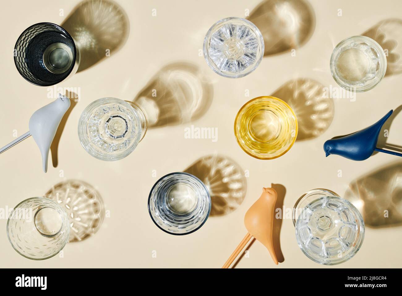 Plusieurs verres contenant de l'eau propre, leurs ombres et trois oiseaux en plastique de couleur bleue, blanche et beige sur fond pastel Banque D'Images
