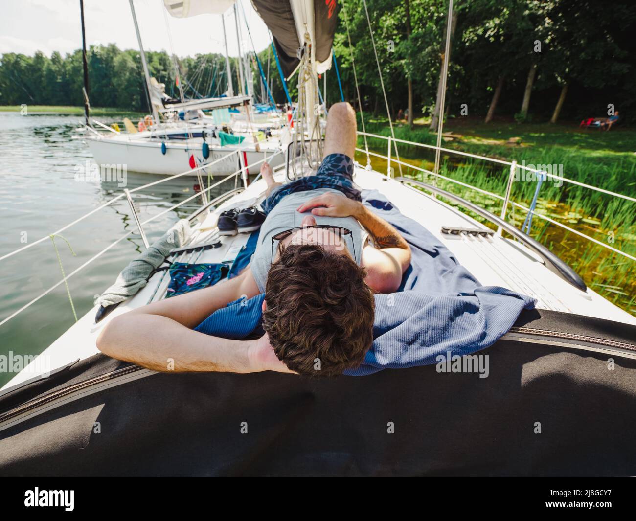 homme adulte se posant sur un pont de voilier et prenant un bain de soleil pendant ses vacances dans une journée ensoleillée d'été. Yacht amarré sur une jetée en bois sur un bord de lac Banque D'Images