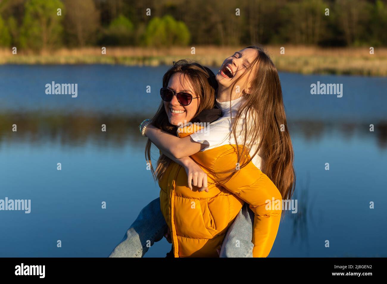Une mère ludique qui donne à sa fille une balade à dos pigeeux au bord du lac de printemps. Rire et paraître heureux. Printemps en arrière-plan du lac. Gros plan. Banque D'Images