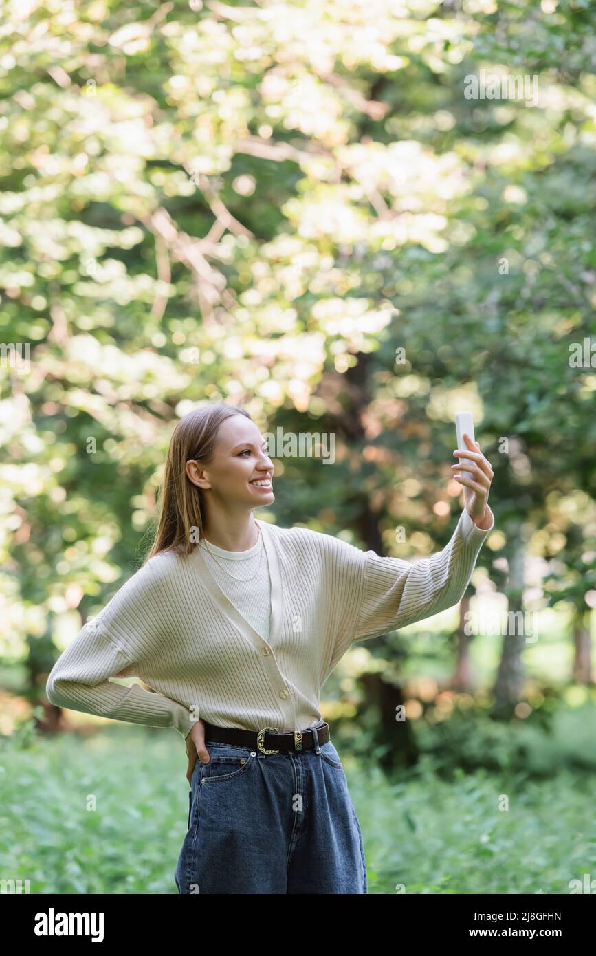 jeune femme heureuse avec un smartphone debout avec la main sur la hanche dans le parc Banque D'Images