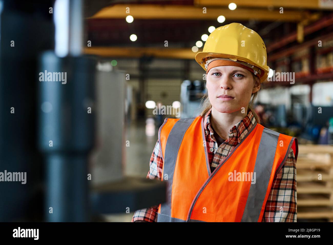 Jeune femme travaillant dans une usine moderne en veste orange et casque de protection regardant la caméra dans un grand atelier ou un entrepôt Banque D'Images