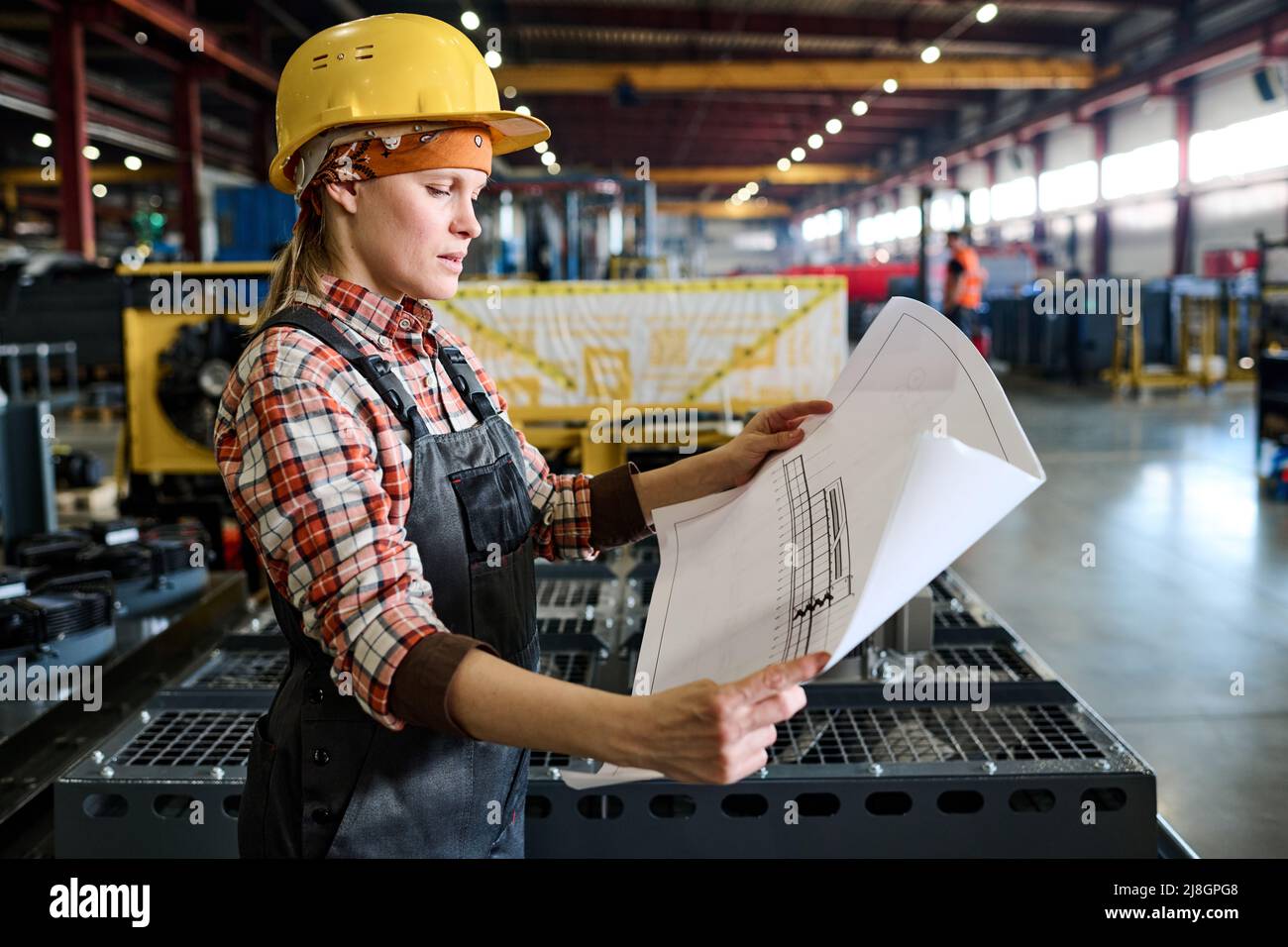 Vue latérale d'une jeune femme ingénieur en vêtements de travail tenant un plan non roulé avec un croquis de construction ou de pièce de machine Banque D'Images