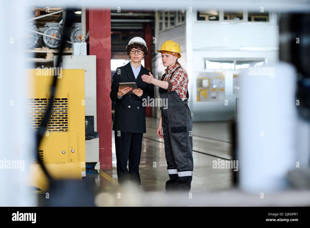 Un jeune ingénieur confiant pointe vers de nouveaux équipements industriels lors d'une conversation avec un collègue ou un responsable avec une tablette lors d'une réunion en usine Banque D'Images