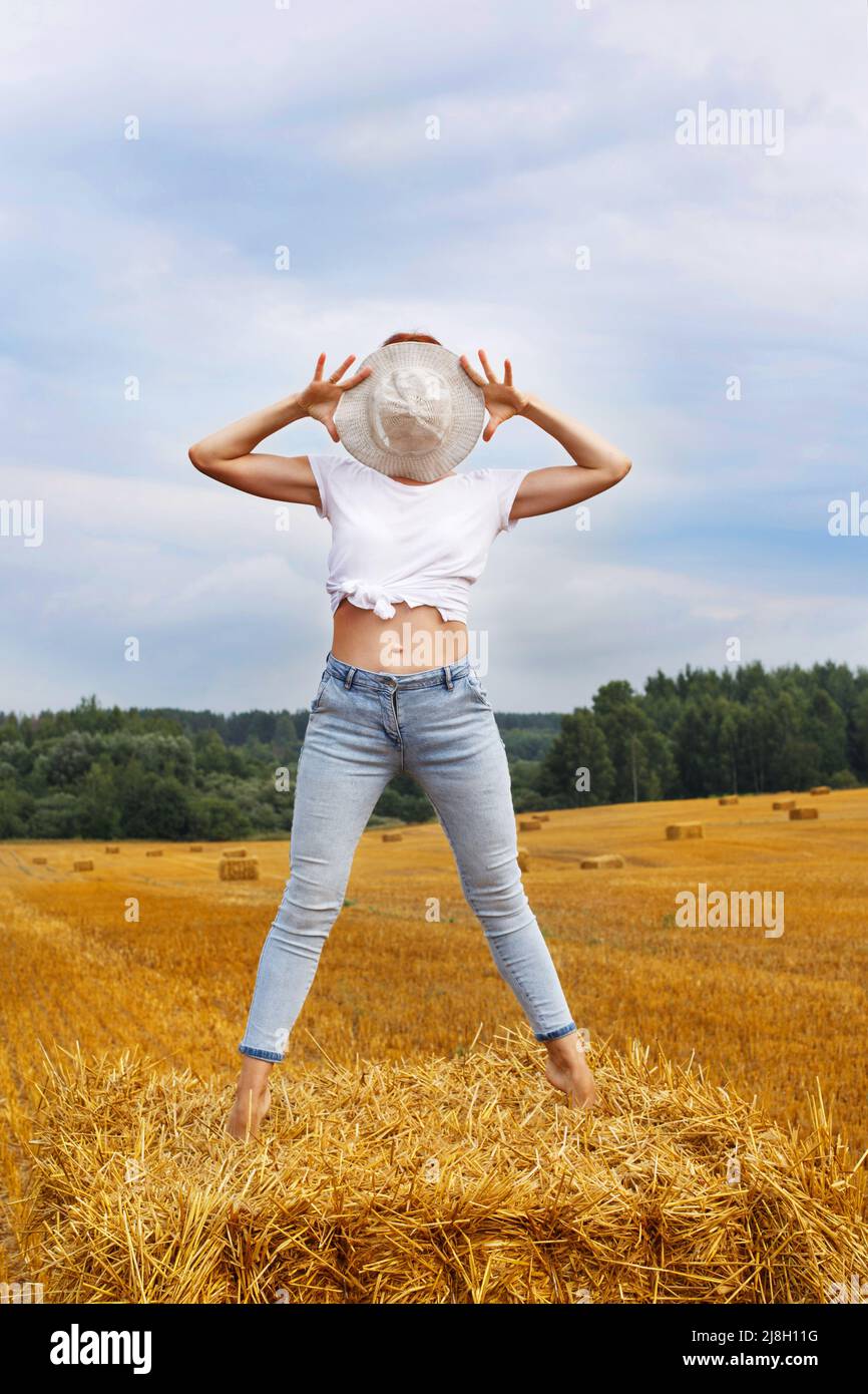 une fille pieds nus en chapeau de paille se tient sur une botte de foin sur une balle dans le champ agricole après la récolte. Banque D'Images