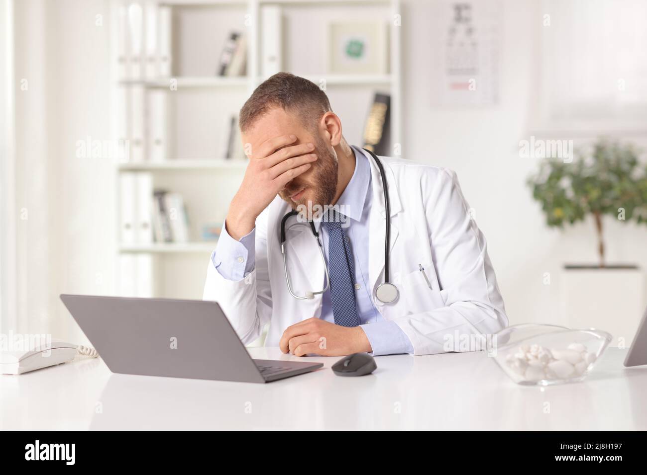 Un homme stressé médecin assis dans un bureau avec un ordinateur portable et tenant sa tête Banque D'Images