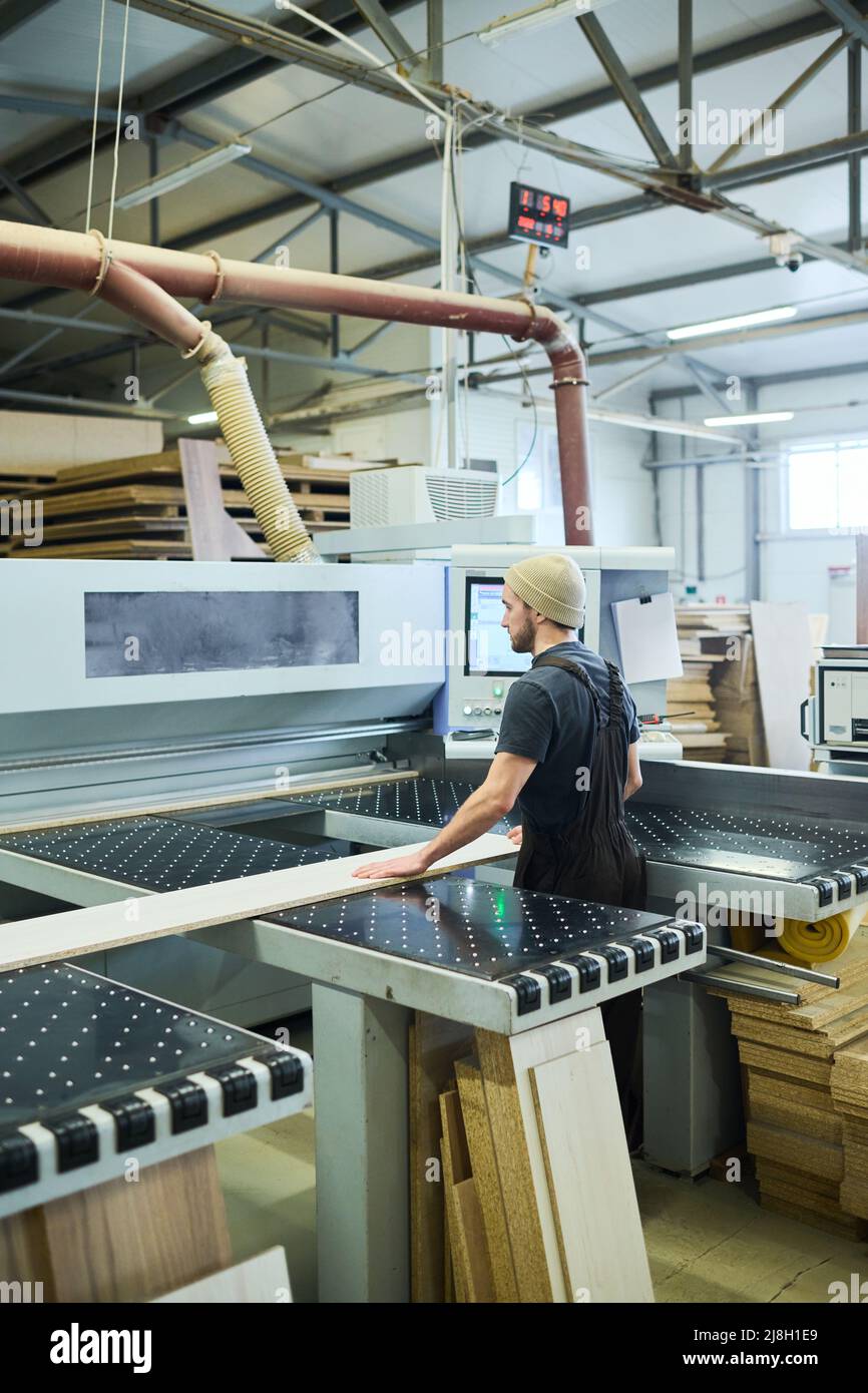 Vue arrière d'un jeune travailleur en position debout sur une grande machine et travaillant avec des matériaux en bois, il contrôle le processus Banque D'Images