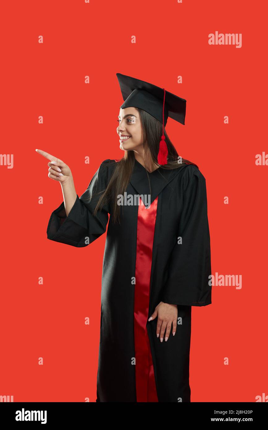 Vue latérale de jolie fille debout, souriant, montrant par le doigt.belle jeune femme diplômé de l'université, portant robe de diplômé et de mortarboard. Est Banque D'Images Vue latérale de jolie fille debout, souriant, montrant par le doigt.belle jeune femme diplômé de l'université, portant robe de diplômé et de mortarboard. Est Banque D'Images