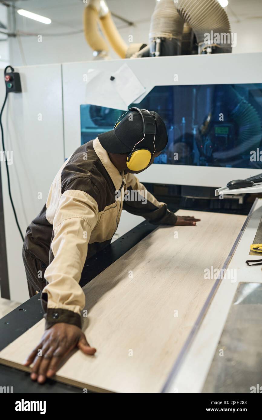 Travailleur africain en casque de protection mettant une planche en bois sur la machine à scier pour fabriquer des meubles à l'usine Banque D'Images