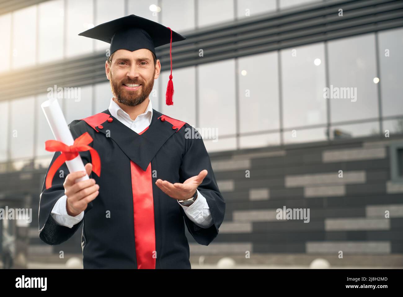 Vue de face d'un beau garçon portant un tableau de bord et une robe de graduation debout, titulaire d'un diplôme. Brunette homme avec barbe diplômé de l'université, smi Banque D'Images Vue de face d'un beau garçon portant un tableau de bord et une robe de graduation debout, titulaire d'un diplôme. Brunette homme avec barbe diplômé de l'université, smi Banque D'Images