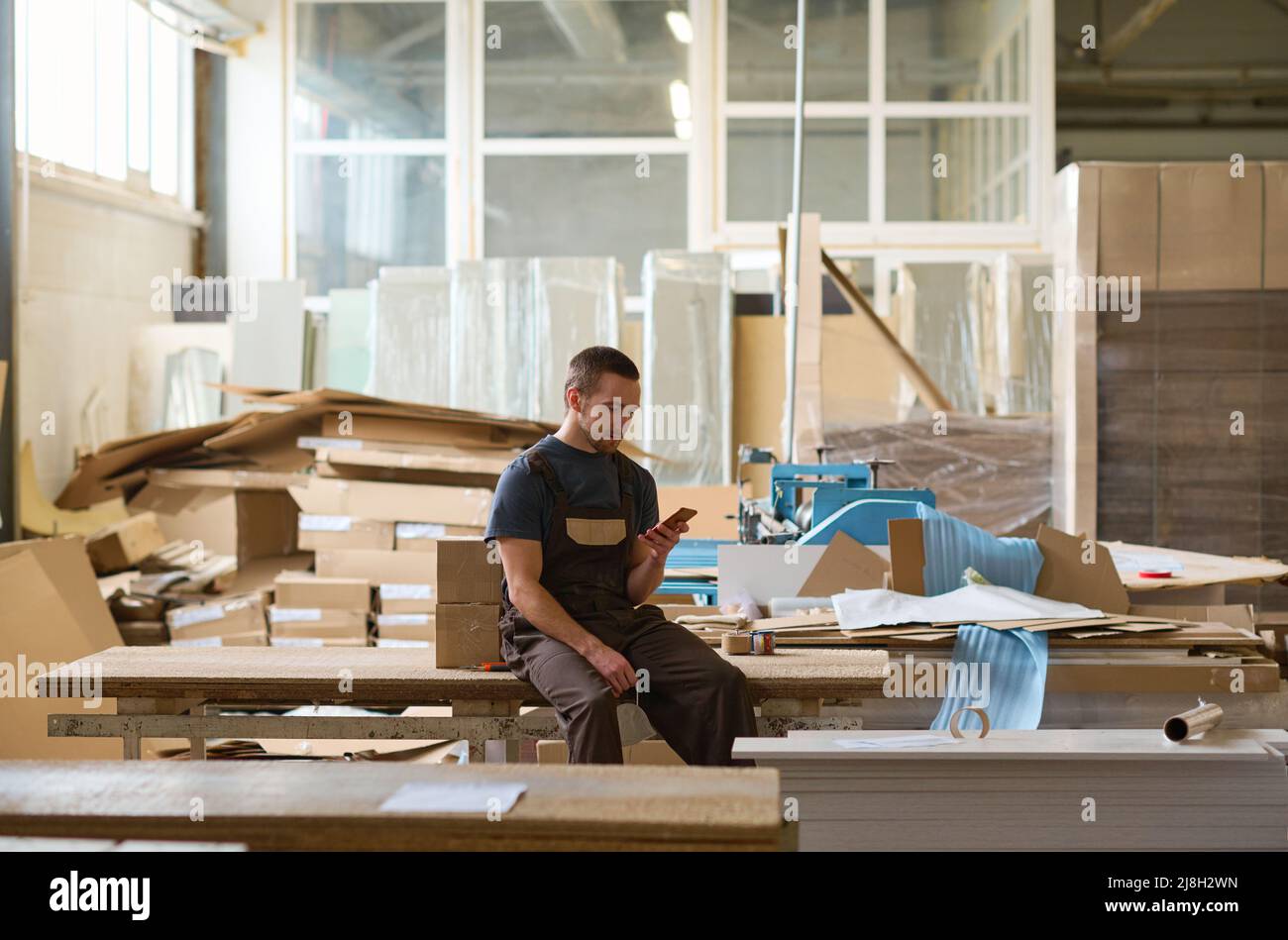 Travailleur manuel assis sur une table et utilisant son téléphone portable pendant sa pause après le travail dans l'entrepôt Banque D'Images