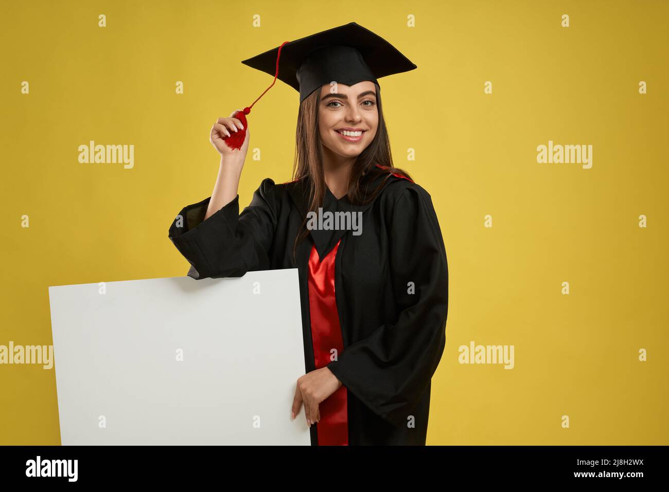 Vue de face d'une charmante femme debout, avec un panneau de mortier. Jolie femme qui obtient son diplôme d'études supérieures, souriant, regardant l'appareil photo. Isolé sur MOC jaune Banque D'Images Vue de face d'une charmante femme debout, avec un panneau de mortier. Jolie femme qui obtient son diplôme d'études supérieures, souriant, regardant l'appareil photo. Isolé sur MOC jaune Banque D'Images