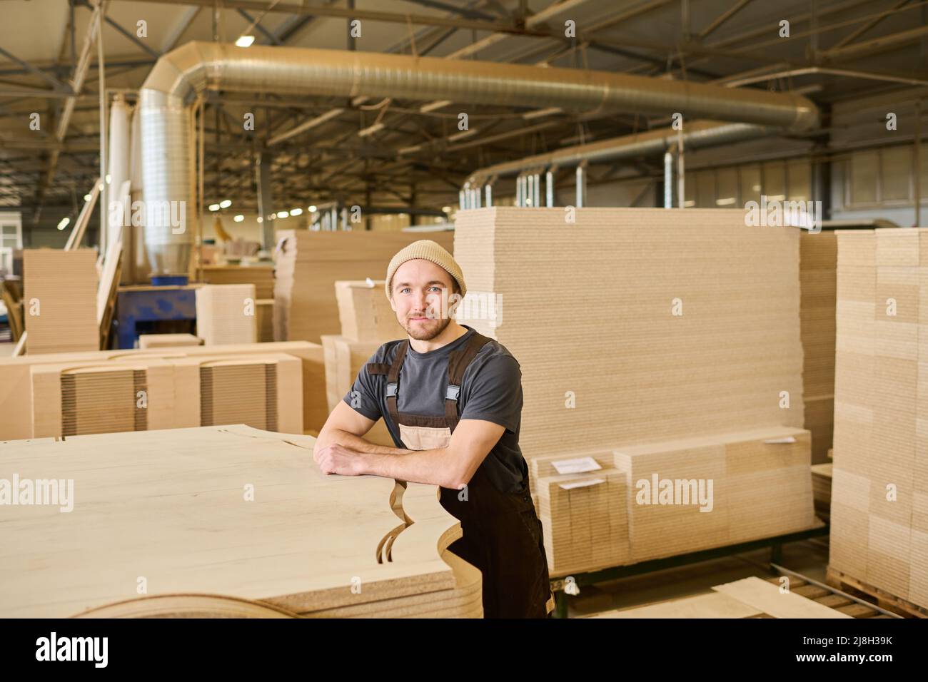 Portrait d'un jeune travailleur dans une combinaison regardant la caméra se tenant à l'entrepôt près des produits en bois Banque D'Images