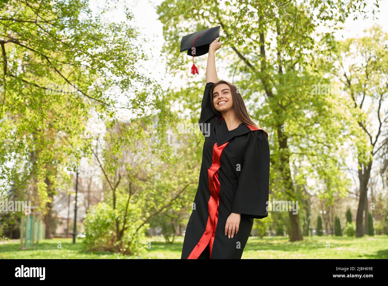 Vue de face de la jolie fille portant une robe de troisième cycle levant le mortaboard. Brunette jeune femme diplômée d'université, université, debout avec fermé e Banque D'Images Vue de face de la jolie fille portant une robe de troisième cycle levant le mortaboard. Brunette jeune femme diplômée d'université, université, debout avec fermé e Banque D'Images