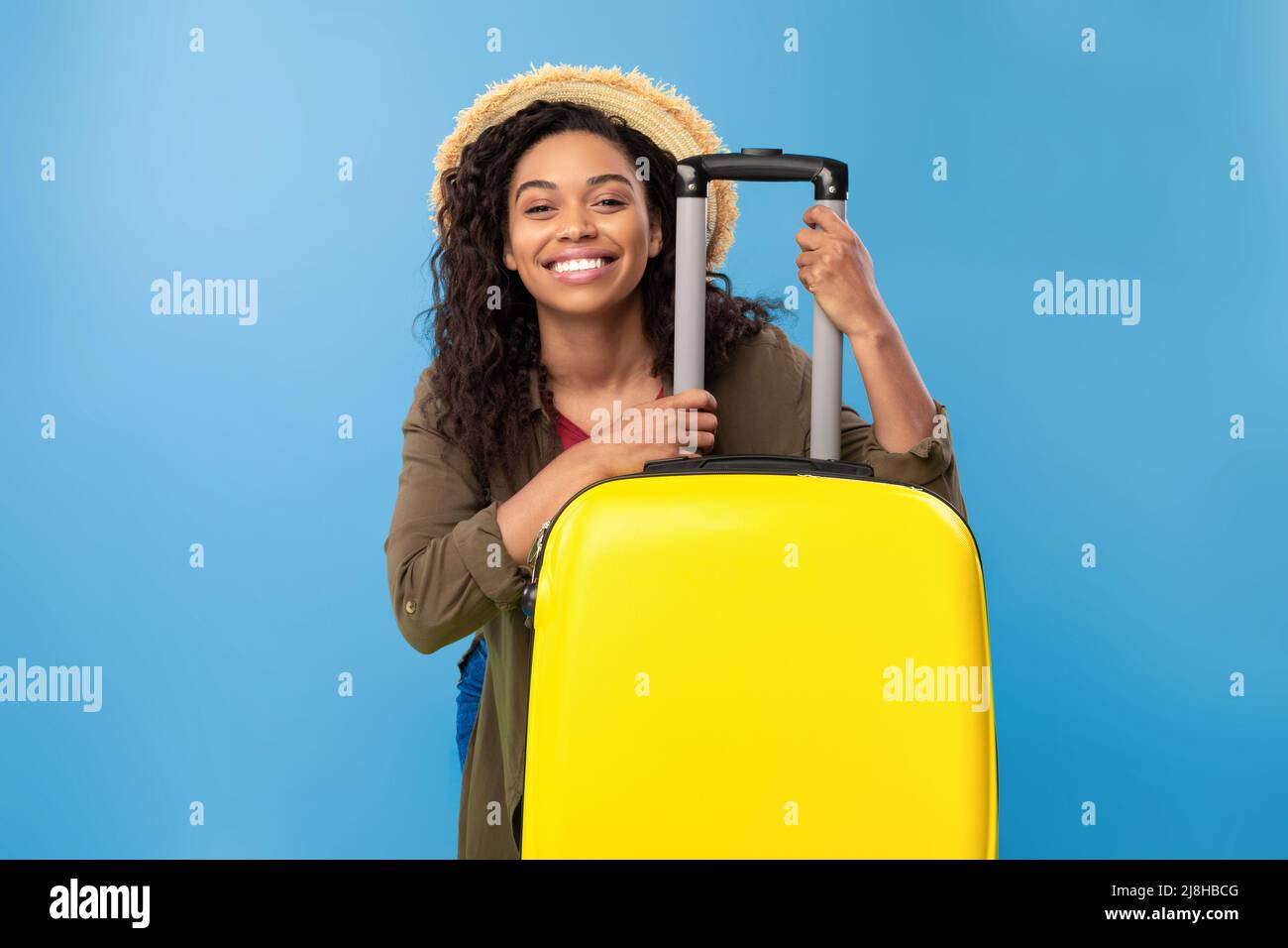 Jeune femme afro-américaine souriante assise derrière une valise jaune vif, en vacances d'été, en voyage à l'étranger Banque D'Images
