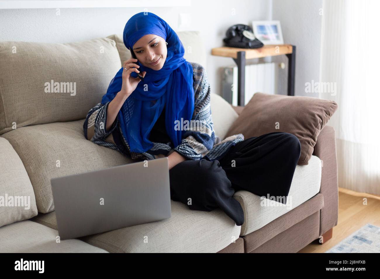 Jeune femme musulmane travaillant à domicile. Elle utilise un ordinateur portable tout en parlant au téléphone. Banque D'Images
