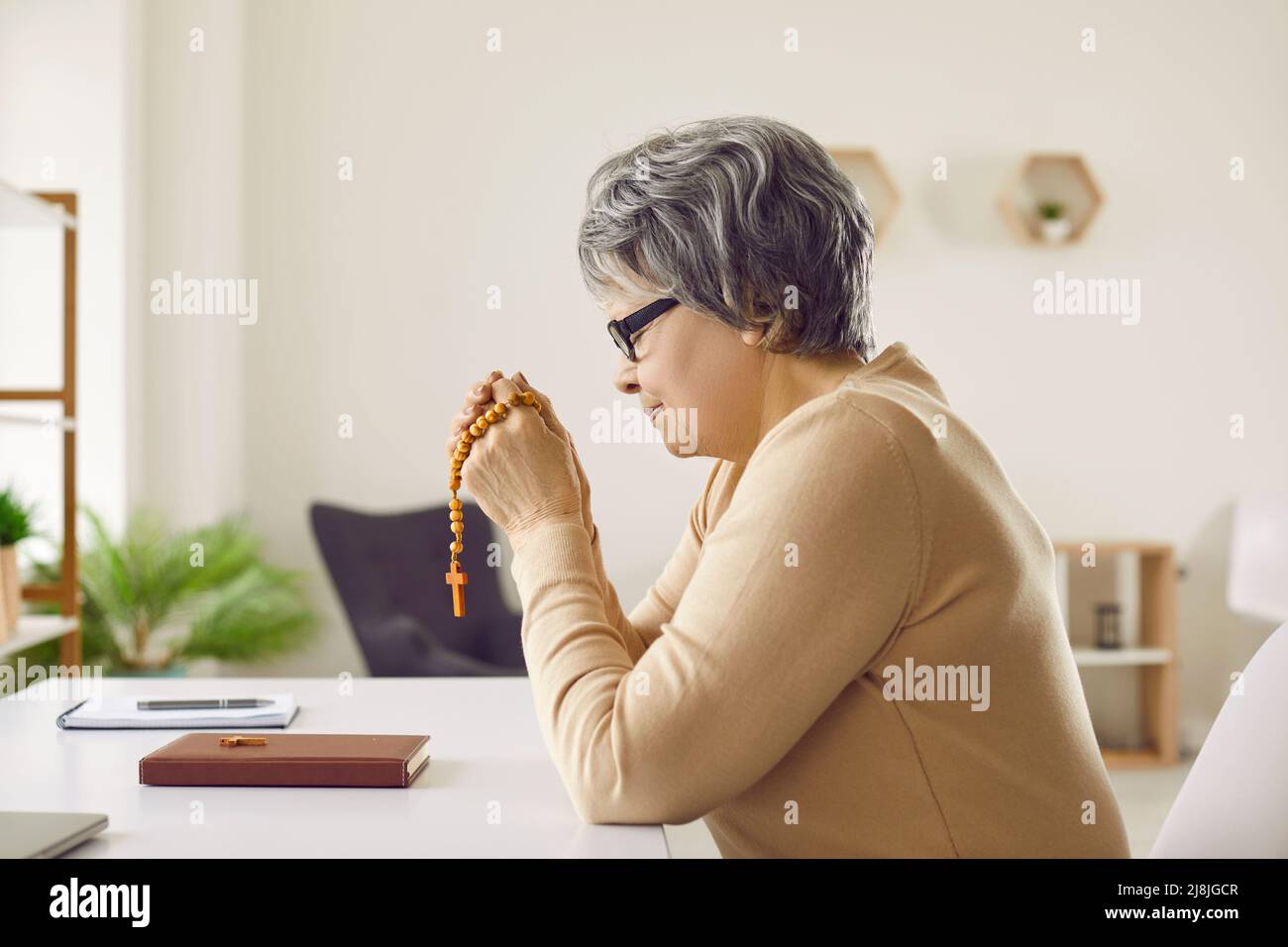 Femme âgée qui appartient à l'Église chrétienne catholique, assise à son bureau et priant Banque D'Images