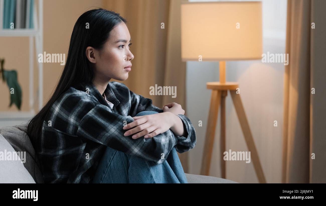 Vue latérale une femme asiatique triste assise sur un canapé à la maison se sentant incertaine d'une décision difficile malheureux un mauvais sentiment mental des ennuis assis sur un canapé. Enfoncé Banque D'Images