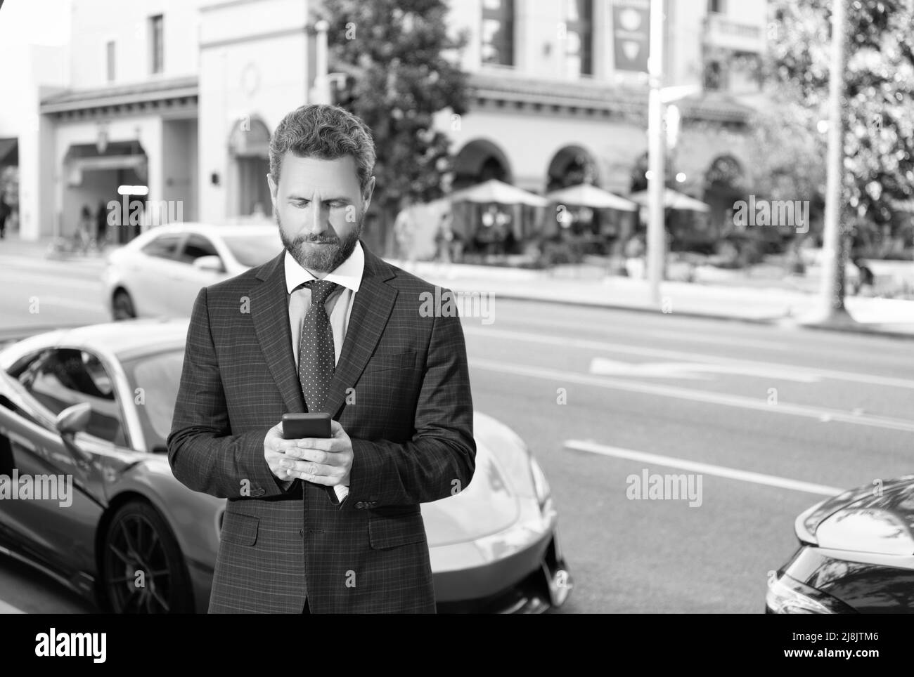 beau homme d'affaires bavardant sur smartphone, à côté d'une voiture de luxe en plein air. voiture disponible. Banque D'Images