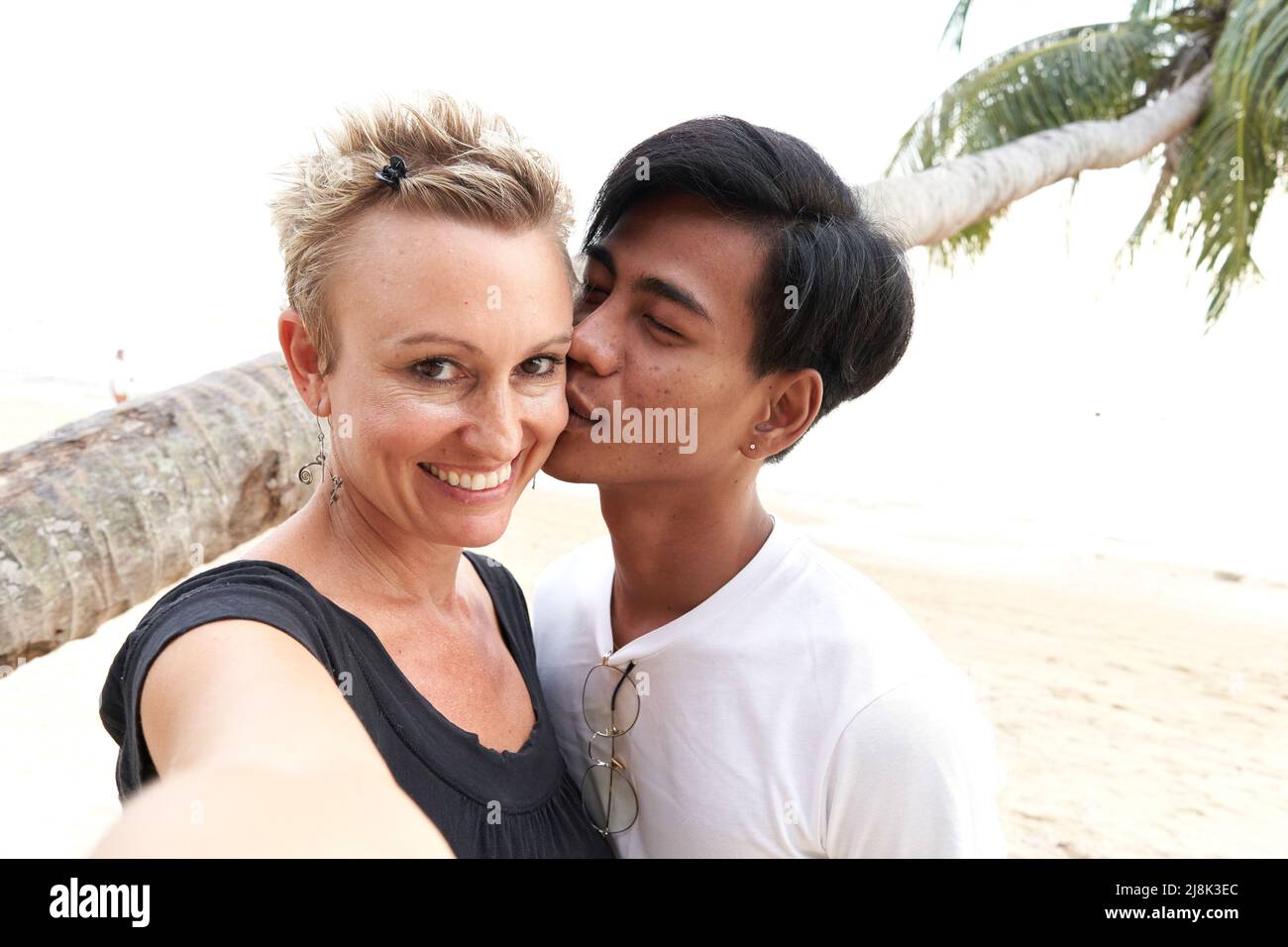 Couple multiculturel embrassant tout en prenant un selfie sur la plage pendant le coucher du soleil Banque D'Images