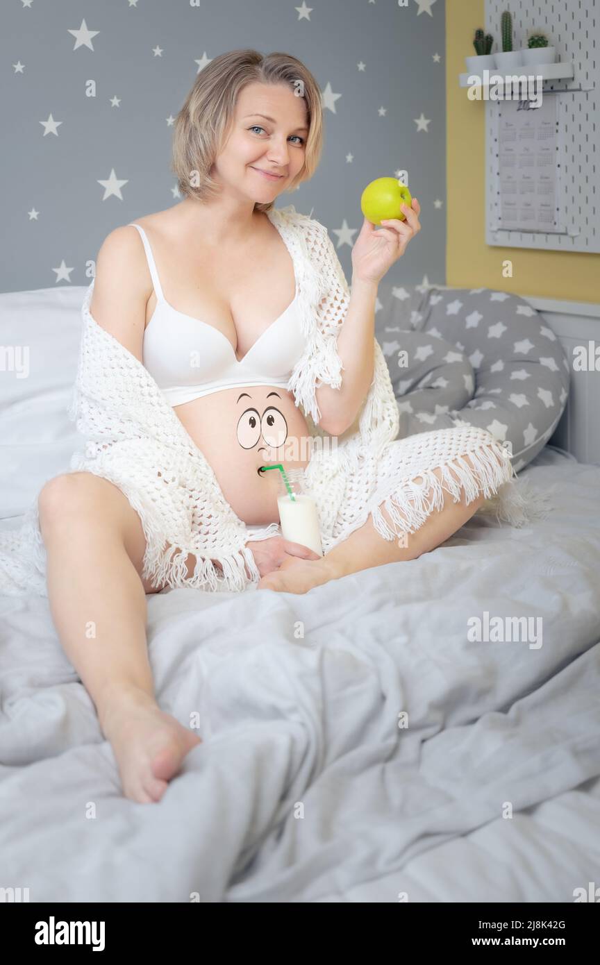 Portrait d'une belle femme enceinte avec des yeux de dessin animé peints sur son ventre, assis sur un lit avec une pomme verte et une bouteille de lait. Une alimentation saine. Banque D'Images