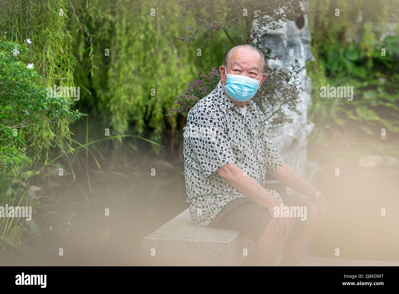 Homme asiatique senior portant un masque, assis sur un banc. Jardin extérieur. Banque D'Images