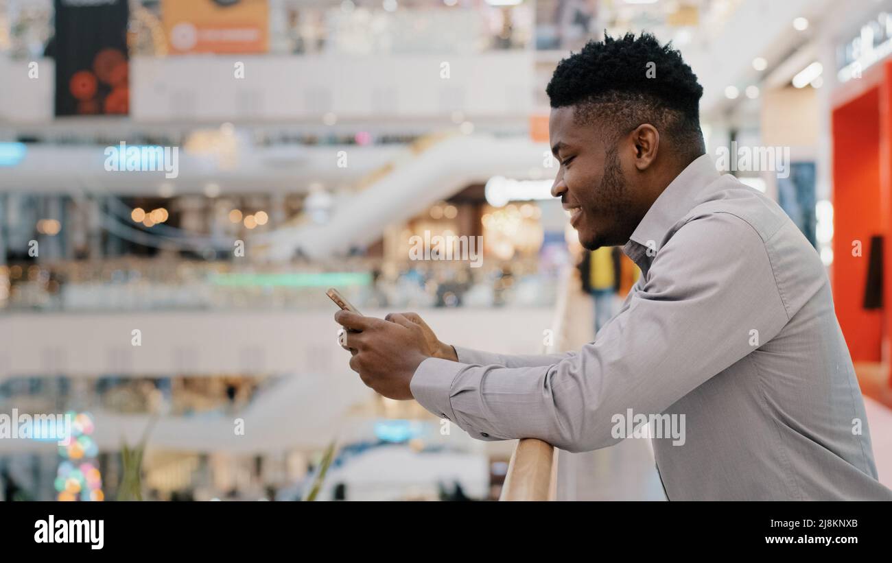 Jeune afro-américain qui se trouve dans un centre commercial et qui passe du temps libre sur Internet sur les réseaux sociaux sur smartphone en utilisant le service virtuel agréable Banque D'Images