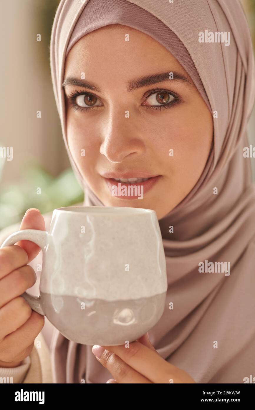 Portrait vertical en gros plan de jeune adulte attrayante Moyen-Orient femme portant le hijab rose pastel élégant boire du thé regardant l'appareil photo Banque D'Images