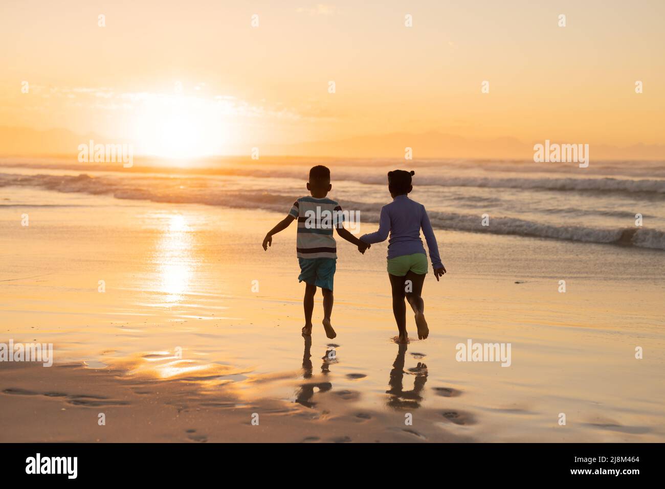 Vue arrière des frères et sœurs afro-américains qui se tiennent les mains et qui couent sur la plage contre le ciel au coucher du soleil Banque D'Images