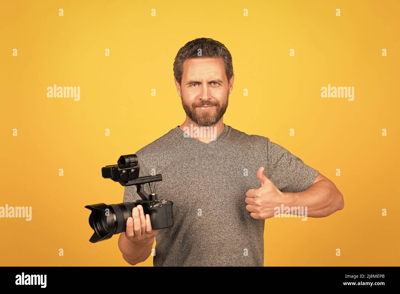 homme barbu plein de gaieté vidéaste pour vlog avec un caméscope montrant le pouce vers le haut, filmant Banque D'Images