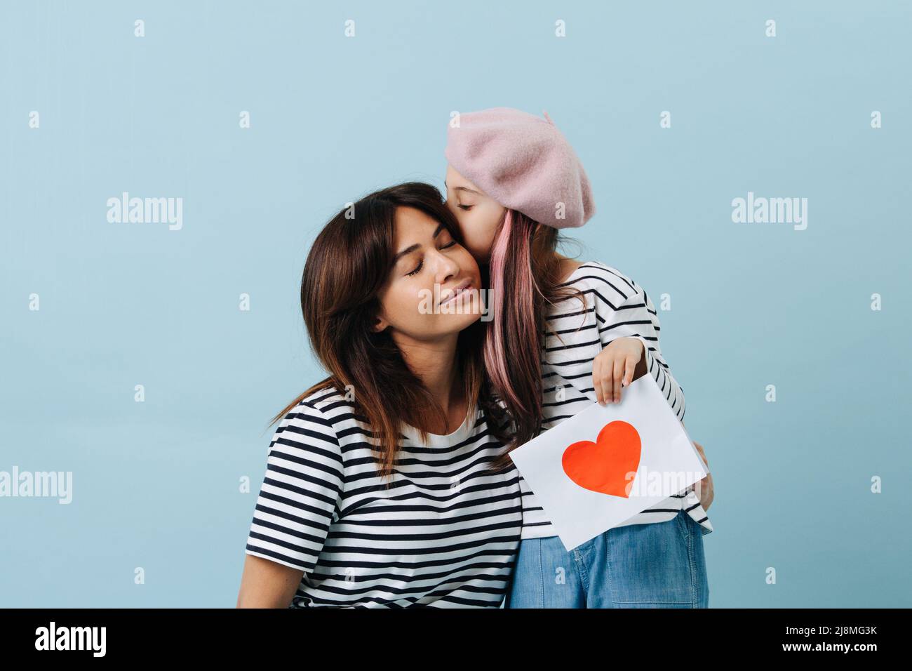 Tween fille en béret français embrassant sa mère, elle tient une feuille de papier avec le coeur écarlate dessus. Sur fond bleu. Banque D'Images