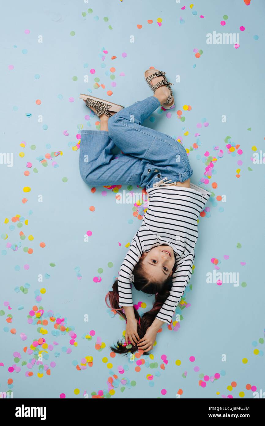 Image à l'envers d'une jolie fille souriante, couchée sur un sol bleu. Entouré de confettis colorés. Banque D'Images