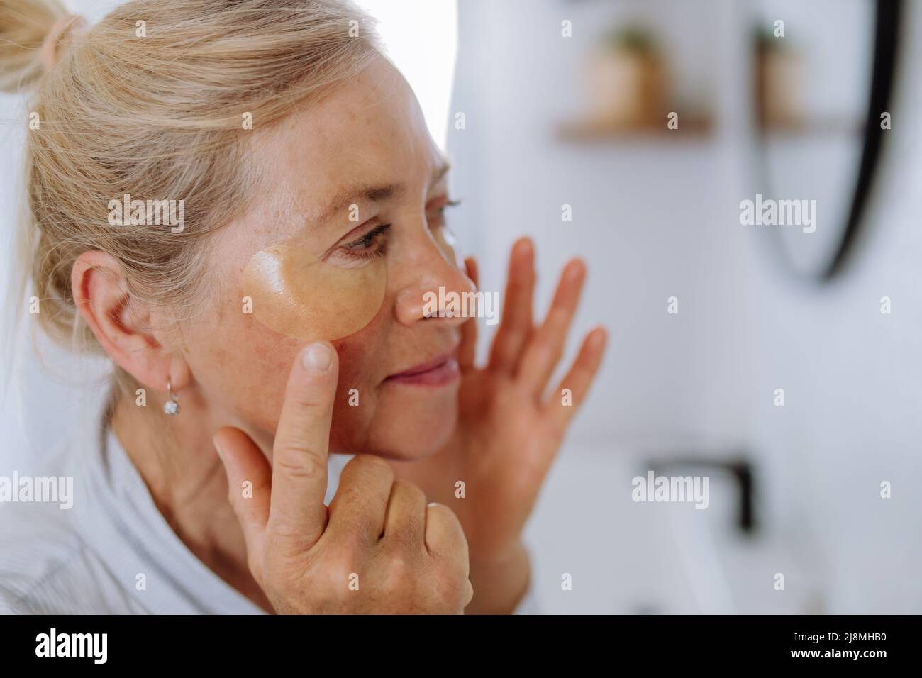 Belle femme âgée en peignoir, application de cellules oculaires pour le puffiness tout en regardant dans le miroir Banque D'Images