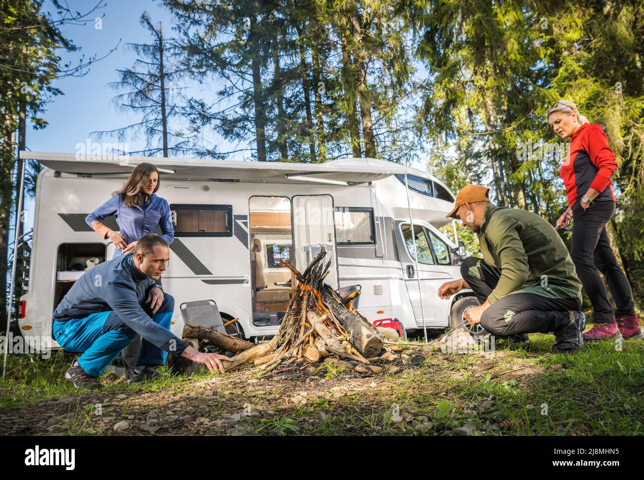 Vacances en voiture de camping avec des amis. Deux couples caucasiens suspendus autour de Campfire à côté de leur camping-car Van. Camping sec dans une forêt. Banque D'Images