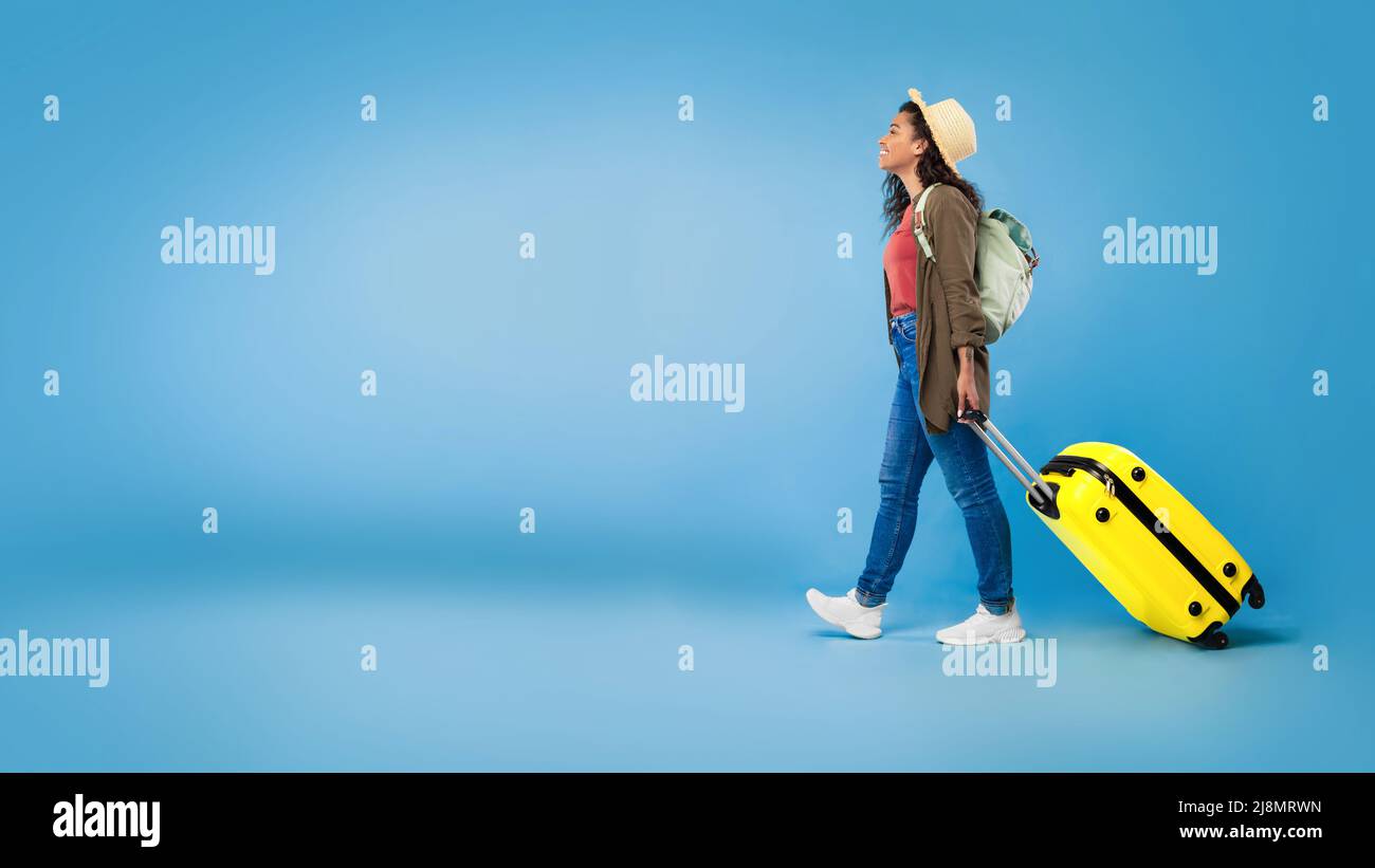 Bonne jeune femme noire marchant avec une valise, voyageant à l'étranger, ayant des vacances d'été sur fond bleu, espace de copie Banque D'Images