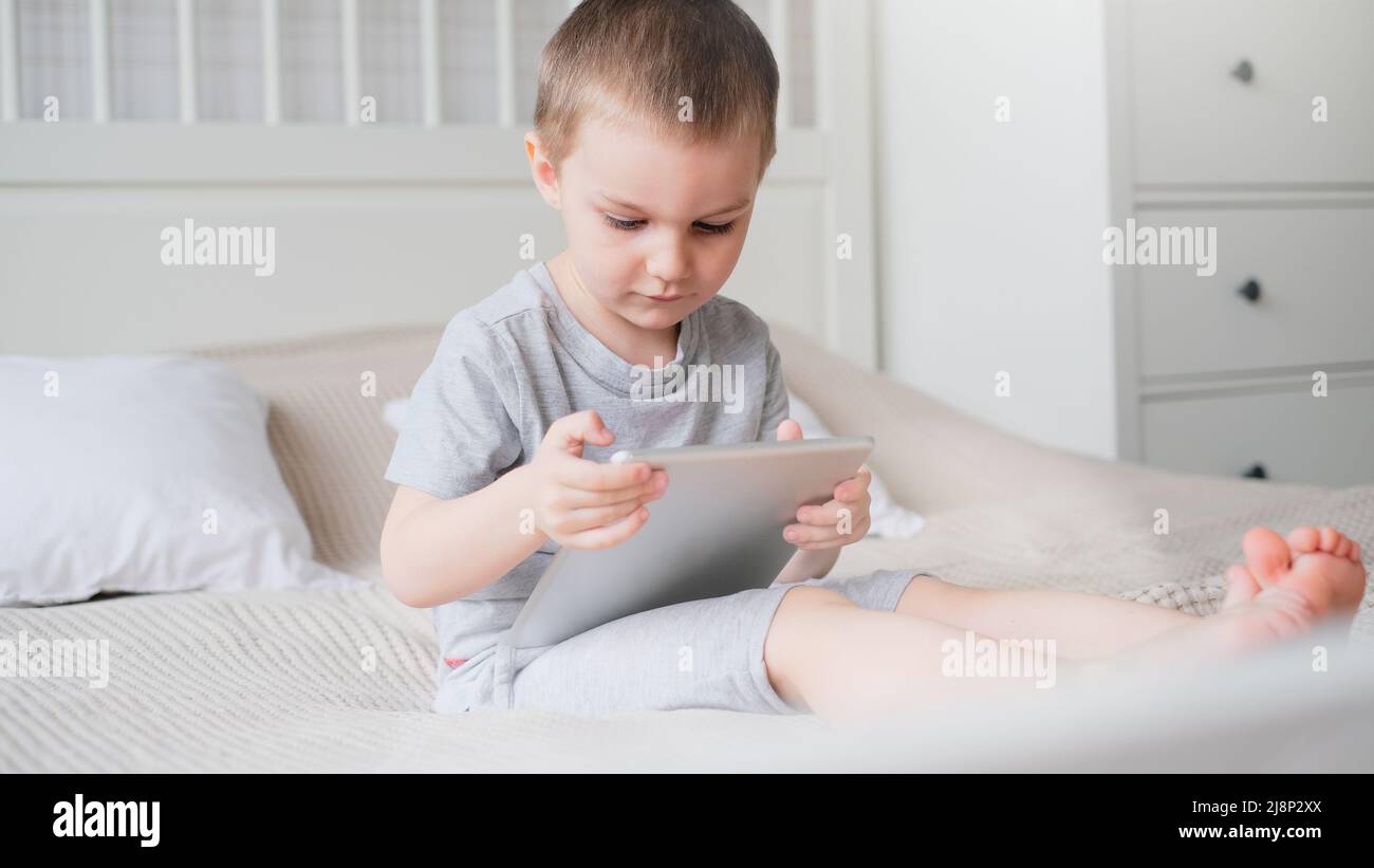 Un petit garçon caucasien dans une pièce lumineuse dans un T-shirt gris joue avec une tablette tout en étant assis sur le canapé. Banque D'Images