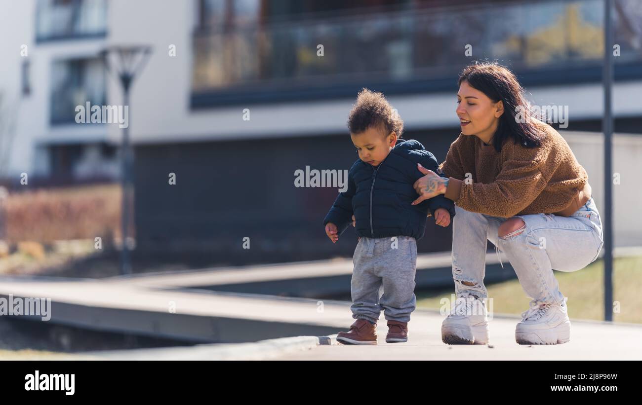 Printemps. Jeune mère brune adulte tenant son petit fils multiracial sur la chaussée. Famille profitant du temps ensoleillé. Photo de haute qualité Banque D'Images