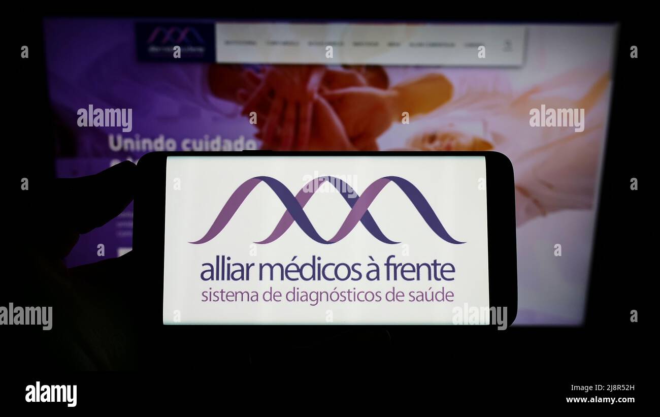 Personne tenant un téléphone portable avec le logo de la société brésilienne de soins de santé Grupo Alliar à l'écran en face de la page Web d'affaires. Mise au point sur l'affichage du téléphone. Banque D'Images