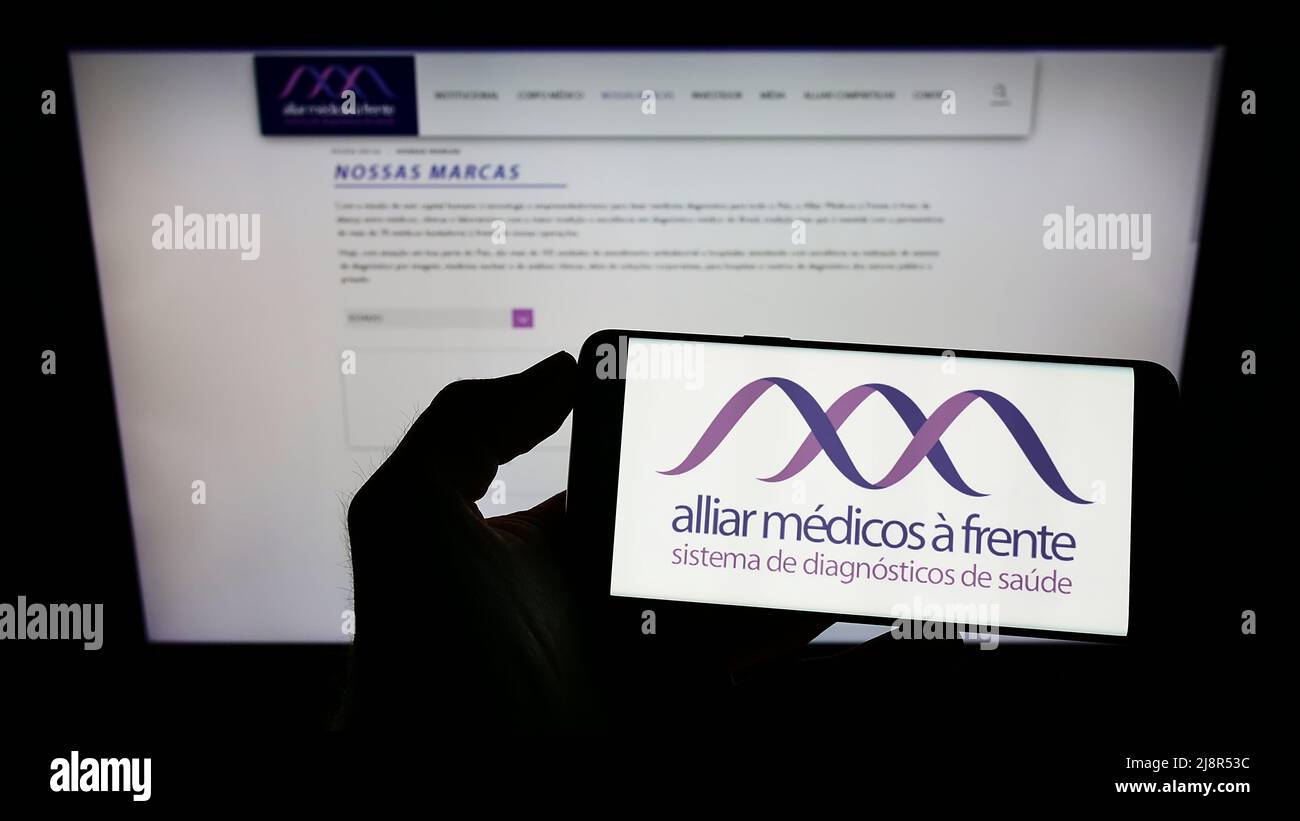 Personne tenant un téléphone portable avec le logo de la société brésilienne de soins de santé Grupo Alliar à l'écran en face de la page Web d'affaires. Mise au point sur l'affichage du téléphone. Banque D'Images