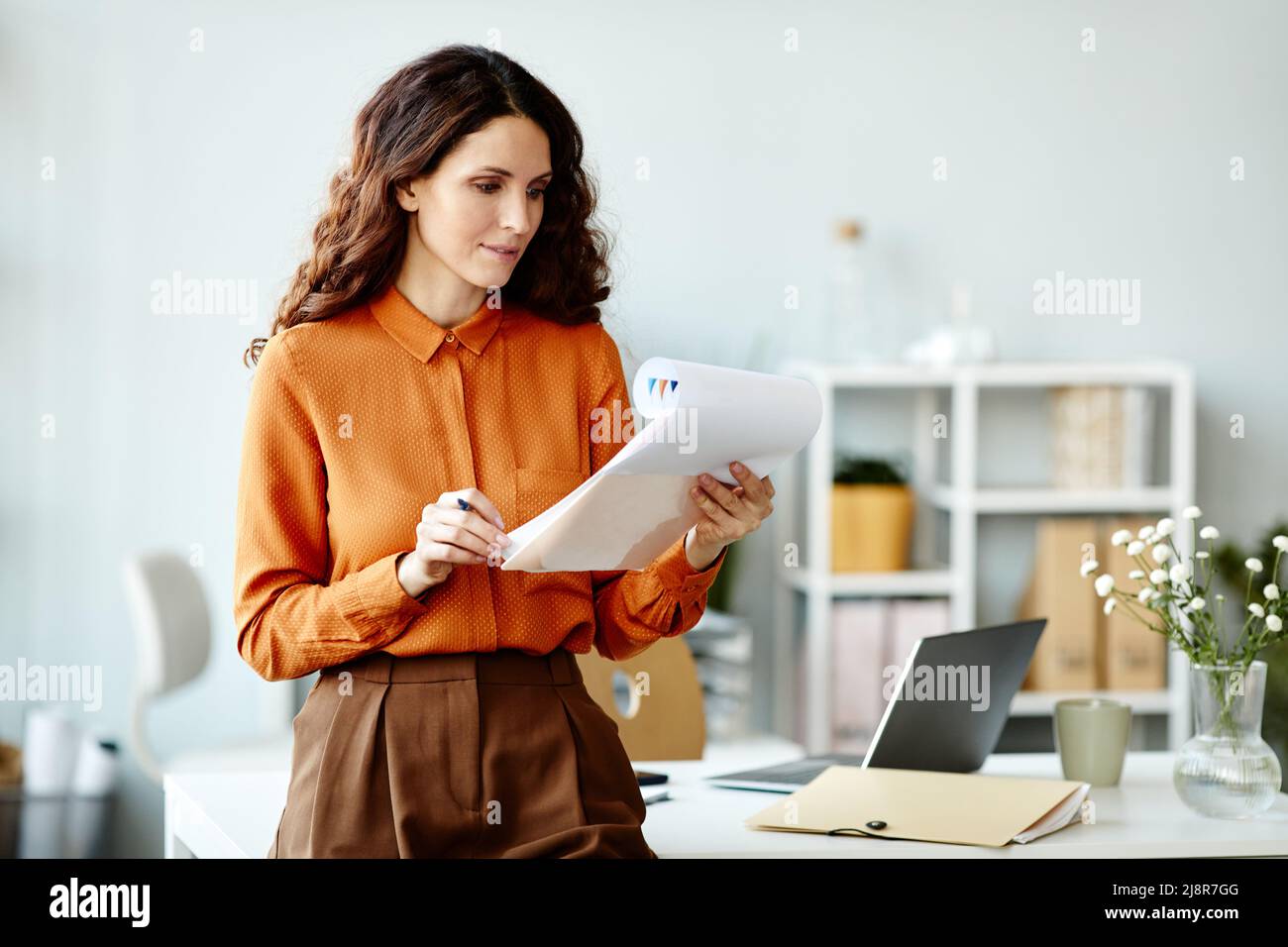 Jeune femme adulte attrayante avec de longs cheveux ondulés debout dans le bureau regardant à travers les documents financiers Banque D'Images