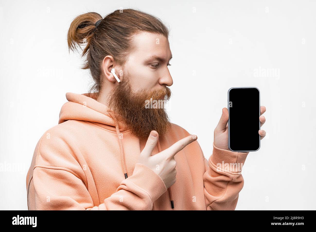 Scandinave beau homme sérieux tenant un téléphone et pointe vers l'écran noir maquette avec casque isolé sur fond gris. Un homme calme avec du gingembre Banque D'Images