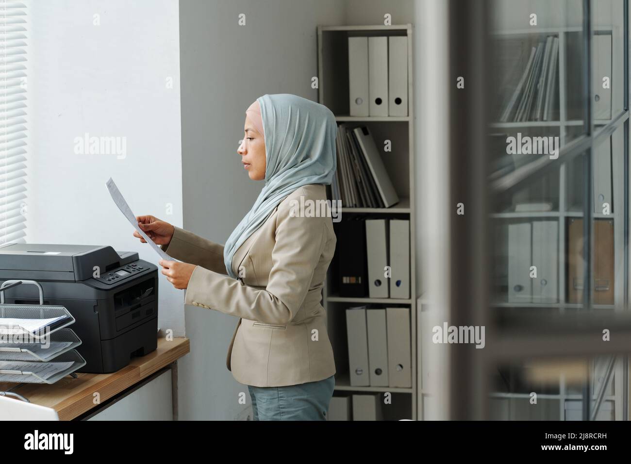 Vue latérale d'une femme musulmane moderne portant le hijab travaillant dans un bureau debout devant l'imprimante vérifiant des copies de document Banque D'Images