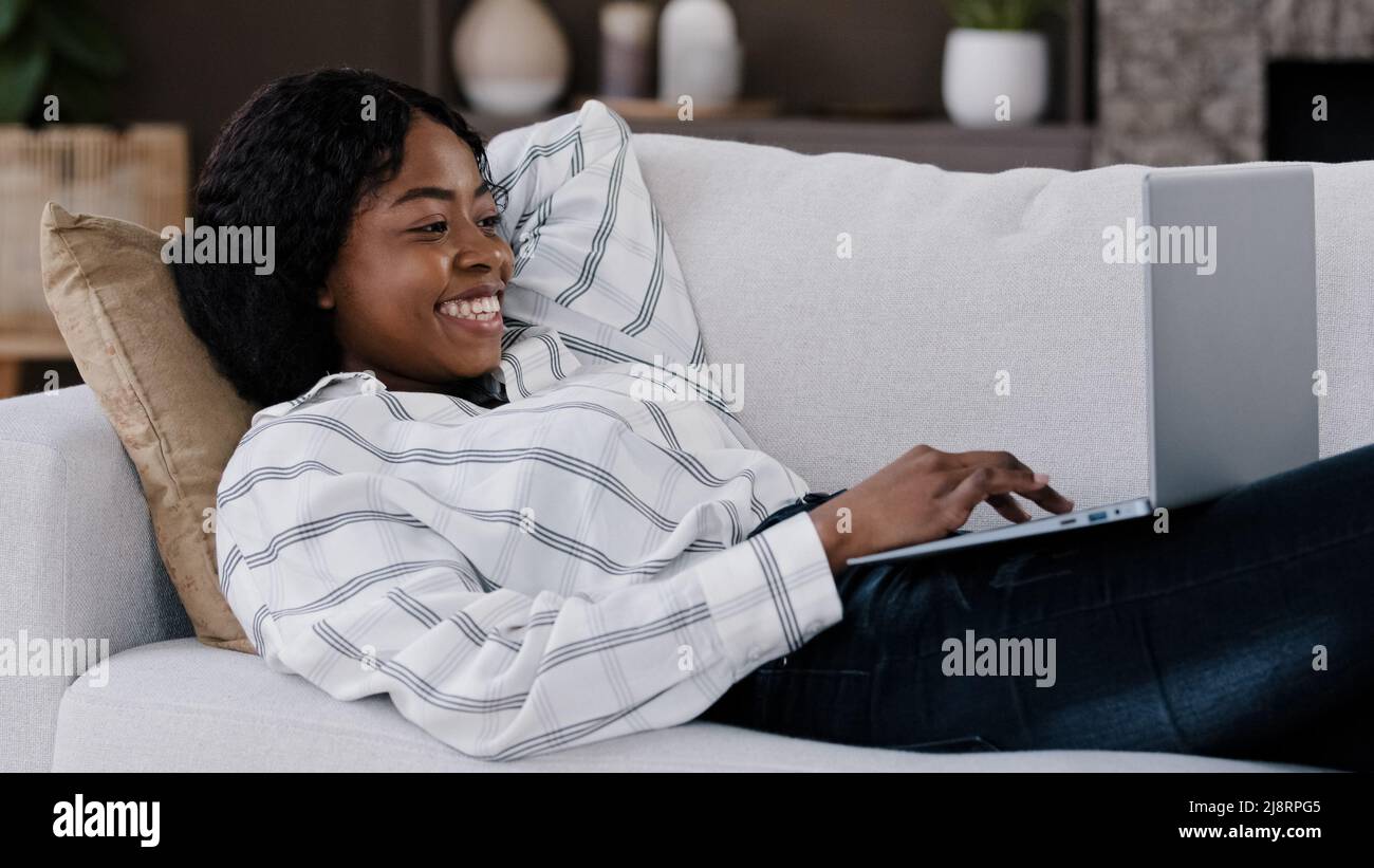 Souriante africaine femme étudiante fille freelancer utilisateur couché sur le canapé en utilisant le PC portable dactylographiant message en ligne chat avec des amis heureux chatter sur le banc d'ordinateur Banque D'Images